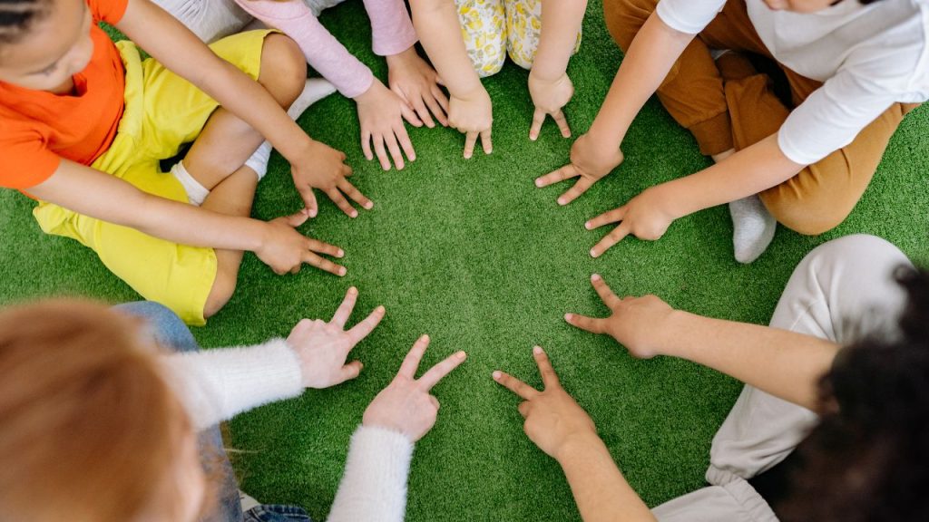 Hands of a diverse group forming a circle on artificial grass, fingers pointing toward the center.
