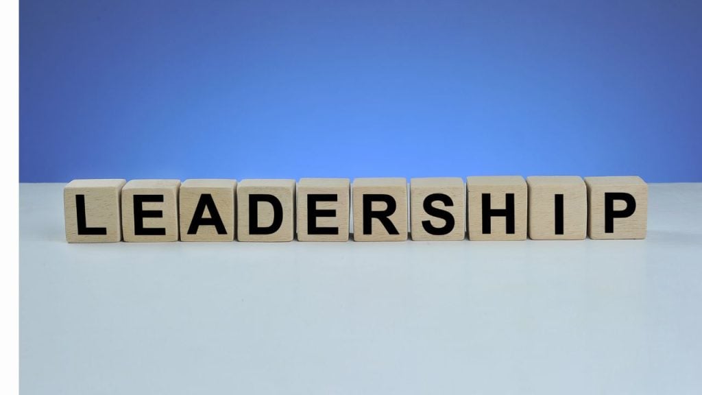 Wooden letter blocks spell 'LEADERSHIP' on a pale desk with a blue gradient background.