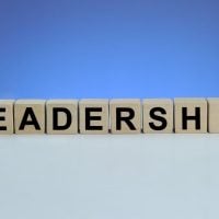 Wooden letter blocks spell 'LEADERSHIP' on a pale desk with a blue gradient background.