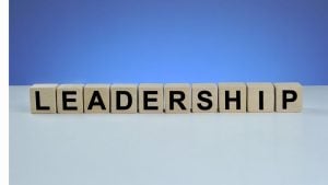 Wooden letter blocks spell 'LEADERSHIP' on a pale desk with a blue gradient background.