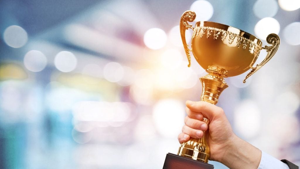 Hand gripping a gold trophy raised in celebration against a bright, blurred background.