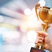 Hand gripping a gold trophy raised in celebration against a bright, blurred background.