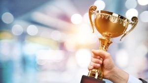Hand gripping a gold trophy raised in celebration against a bright, blurred background.