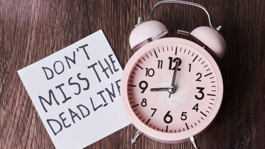 Pink retro alarm clock beside a note reading "Don't miss the deadline" on a dark wooden surface, conveying urgency about deadlines.