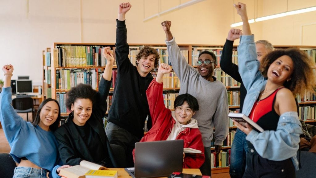 Diverse group of students in a library cheerfully raising their fists around a laptop on a table."
