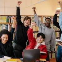 Diverse group of students in a library cheerfully raising their fists around a laptop on a table."