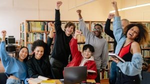 Diverse group of students in a library cheerfully raising their fists around a laptop on a table."