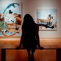 Silhouette of a person seated on a bench in a gallery, facing two large colorful paintings on the wall, with a warm wooden floor and soft overhead lighting.