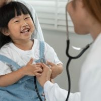 Smiling young girl during a pediatric checkup as a doctor listens to her heart with a stethoscope, with a parent nearby.
