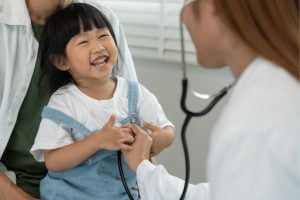 Smiling young girl during a pediatric checkup as a doctor listens to her heart with a stethoscope, with a parent nearby.