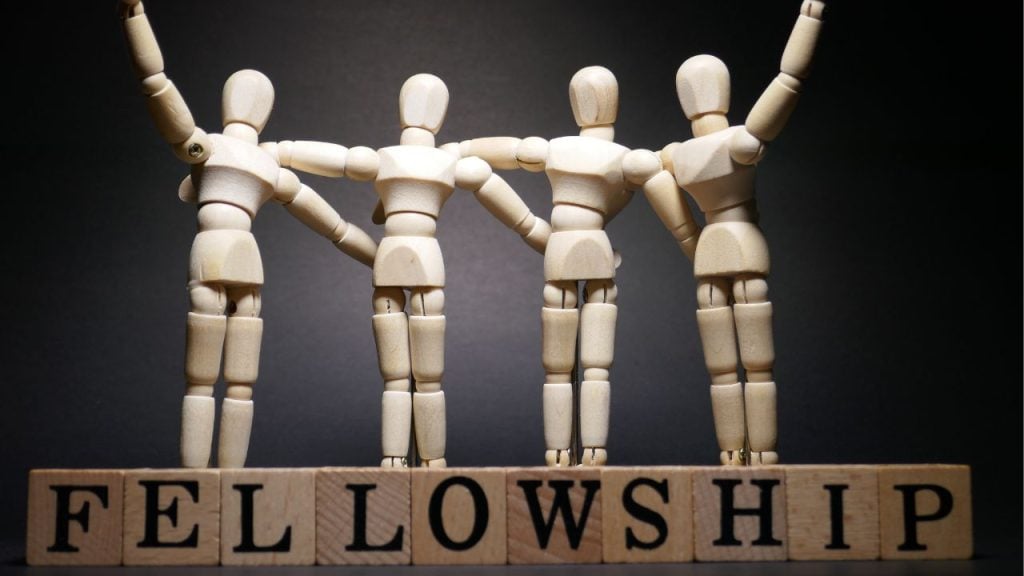 Wooden artist mannequins stand in a row with arms linked, over blocks that spell FELLOWSHIP, symbolizing unity.