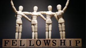 Wooden artist mannequins stand in a row with arms linked, over blocks that spell FELLOWSHIP, symbolizing unity.