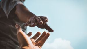 Two hands reaching toward each other against a bright blue sky, suggesting connection or support.