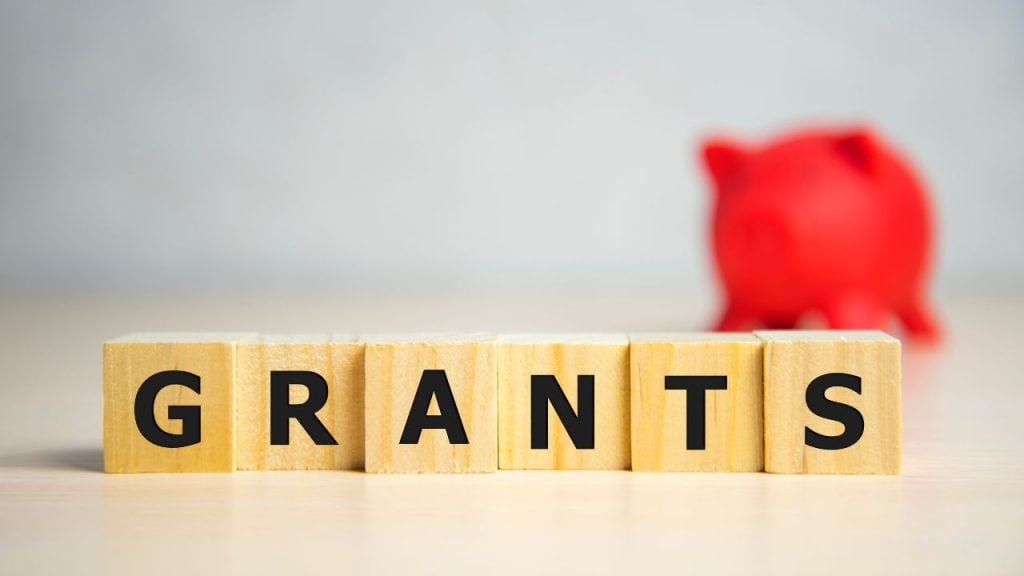 education and sports for children 1 - fundsforNGOs Wooden letter blocks spell 'GRANTS' on a desk, with a red piggy bank blurred in the background.