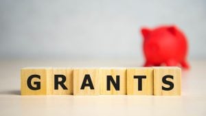Wooden letter blocks spell 'GRANTS' on a desk, with a red piggy bank blurred in the background.