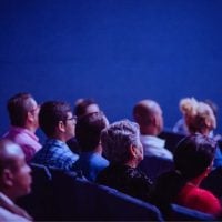 film - fundsforNGOs Audience seated in rows facing a stage in a dark blue-lit theater or conference space.