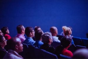 Audience seated in rows facing a stage in a dark blue-lit theater or conference space.