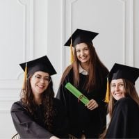 Three graduates in black gowns and mortarboards pose together, one holding a green diploma roll.