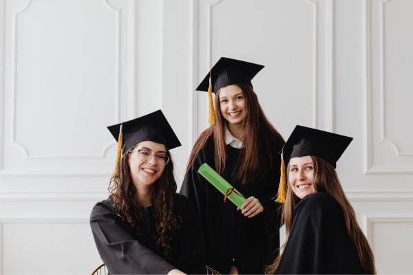 Three graduates in black gowns and mortarboards pose together, one holding a green diploma roll.