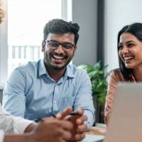 Three colleagues laughing and talking around a laptop in a bright office conference space, mid‑discussion area.