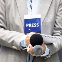 journalist - fundsforNGOs Reporter in a beige suit holds a microphone and notebook with a visible press badge on the chest area lanyard.