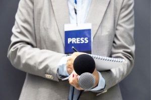 Reporter in a beige suit holds a microphone and notebook with a visible press badge on the chest area lanyard.