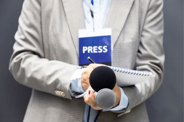 journalist - fundsforNGOs Reporter in a beige suit holds a microphone and notebook with a visible press badge on the chest area lanyard.