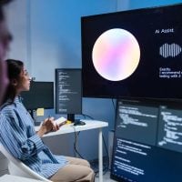 AI - fundsforNGOs Two developers in a modern lab watch a large monitor displaying Ai Assist with a colorful circular graphic and waveform, while code windows are visible on nearby screens.