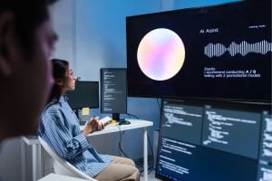 Two developers in a modern lab watch a large monitor displaying Ai Assist with a colorful circular graphic and waveform, while code windows are visible on nearby screens.