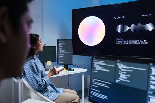 Two developers in a modern lab watch a large monitor displaying Ai Assist with a colorful circular graphic and waveform, while code windows are visible on nearby screens.