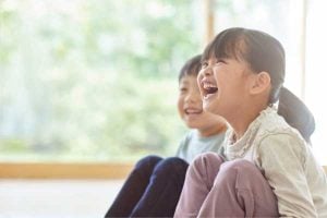 Two children sit on the floor by a window, laughing and enjoying a moment together.