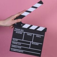 Documentary - fundsforNGOs A hand holds a black clapperboard against a pink background, ready for filming.