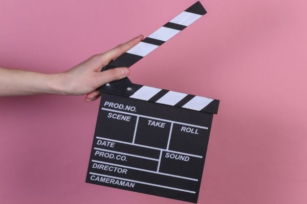 Documentary - fundsforNGOs A hand holds a black clapperboard against a pink background, ready for filming.