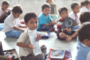 Young boy sits on the classroom floor, holding a small slate and looking at the camera.