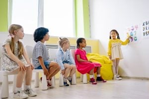 Five children sit on small stools in a bright classroom while one girl in yellow stands by the wall pointing to a chart with letters and numbers.