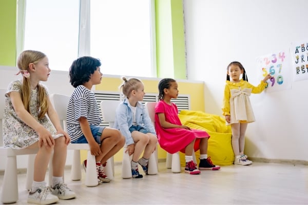 Five children sit on small stools in a bright classroom while one girl in yellow stands by the wall pointing to a chart with letters and numbers.
