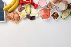 Flat lay of assorted healthy foods lined along a white surface: bananas, avocado, chicken, eggs, dairy, salmon, chickpeas, nuts, chocolate, cheese, and milk.