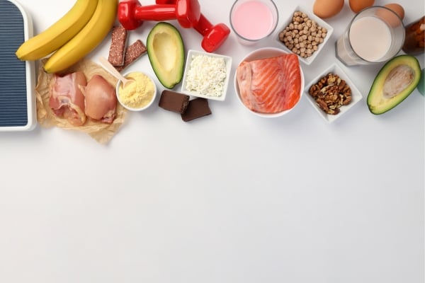 Flat lay of assorted healthy foods lined along a white surface: bananas, avocado, chicken, eggs, dairy, salmon, chickpeas, nuts, chocolate, cheese, and milk.