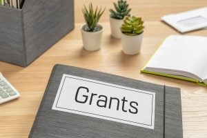 Binder labeled Grants on a wooden desk with small potted plants, an open notebook, and a calculator nearby.