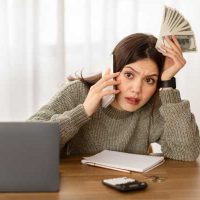 Financial Shock - fundsforNGOs Stressed woman on a call at a desk, holding cash in one hand while talking on her phone, with a laptop and notebook nearby.