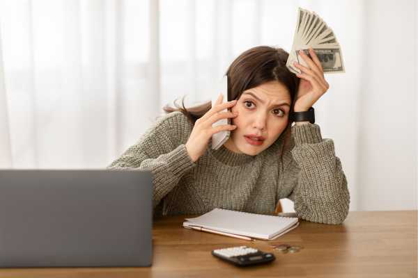 Financial Shock - fundsforNGOs Stressed woman on a call at a desk, holding cash in one hand while talking on her phone, with a laptop and notebook nearby.