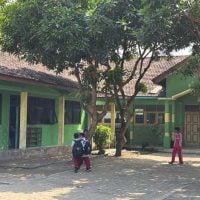 Green Schools - fundsforNGOs School courtyard with lime-green buildings, a large tree in the center, and several children walking around the yard.