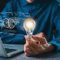 Innovation 1 - fundsforNGOs Person in a blue shirt holds a lit bulb beside a laptop, with a holographic AI brain—symbolizing AI ideas.