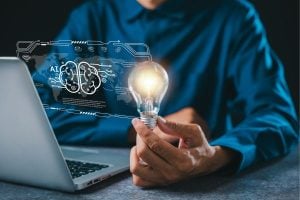 Person in a blue shirt holds a lit bulb beside a laptop, with a holographic AI brain—symbolizing AI ideas.