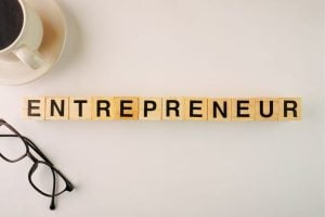 Wooden letter tiles spell 'ENTREPRENEUR' on a light desk with a coffee cup nearby.