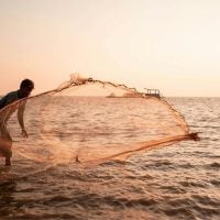 LIVELIHOOD - fundsforNGOs A man casts a circular fishing net into the sea at sunset, the horizon glowing with warm orange light.