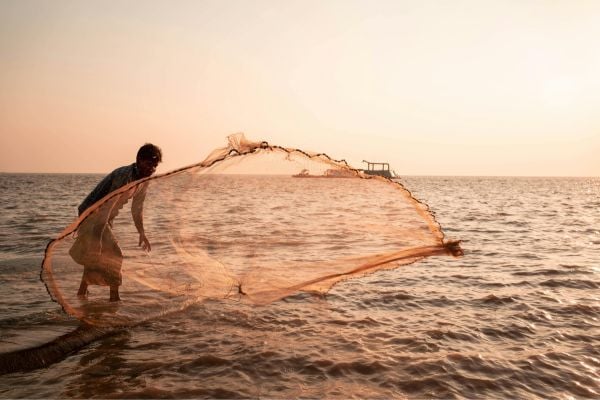 A man casts a circular fishing net into the sea at sunset, the horizon glowing with warm orange light.