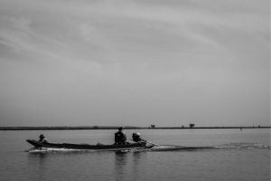 Two people in a small motorboat gliding across calm water with a distant shoreline visible in the background.