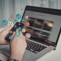 Person typing on a laptop with colorful social media icons and reaction emojis appearing from the keyboard