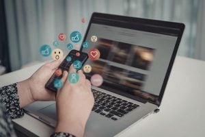 Person typing on a laptop with colorful social media icons and reaction emojis appearing from the keyboard